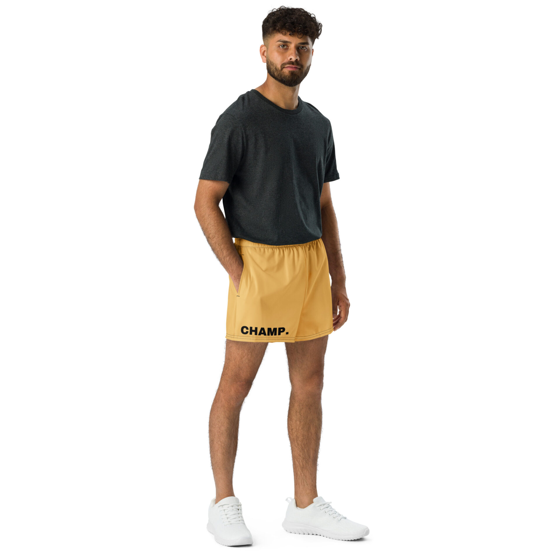 A person with short curly hair stands against a white background, wearing a dark gray t-shirt, CHAMP Golden Athletic Shorts, and white sneakers. They have one hand in their pocket and look directly at the camera. A person with short curly hair stands against a white background, wearing a dark gray t-shirt, CHAMP Golden Athletic Shorts, and white sneakers. They have one hand in their pocket and look directly at the camera.