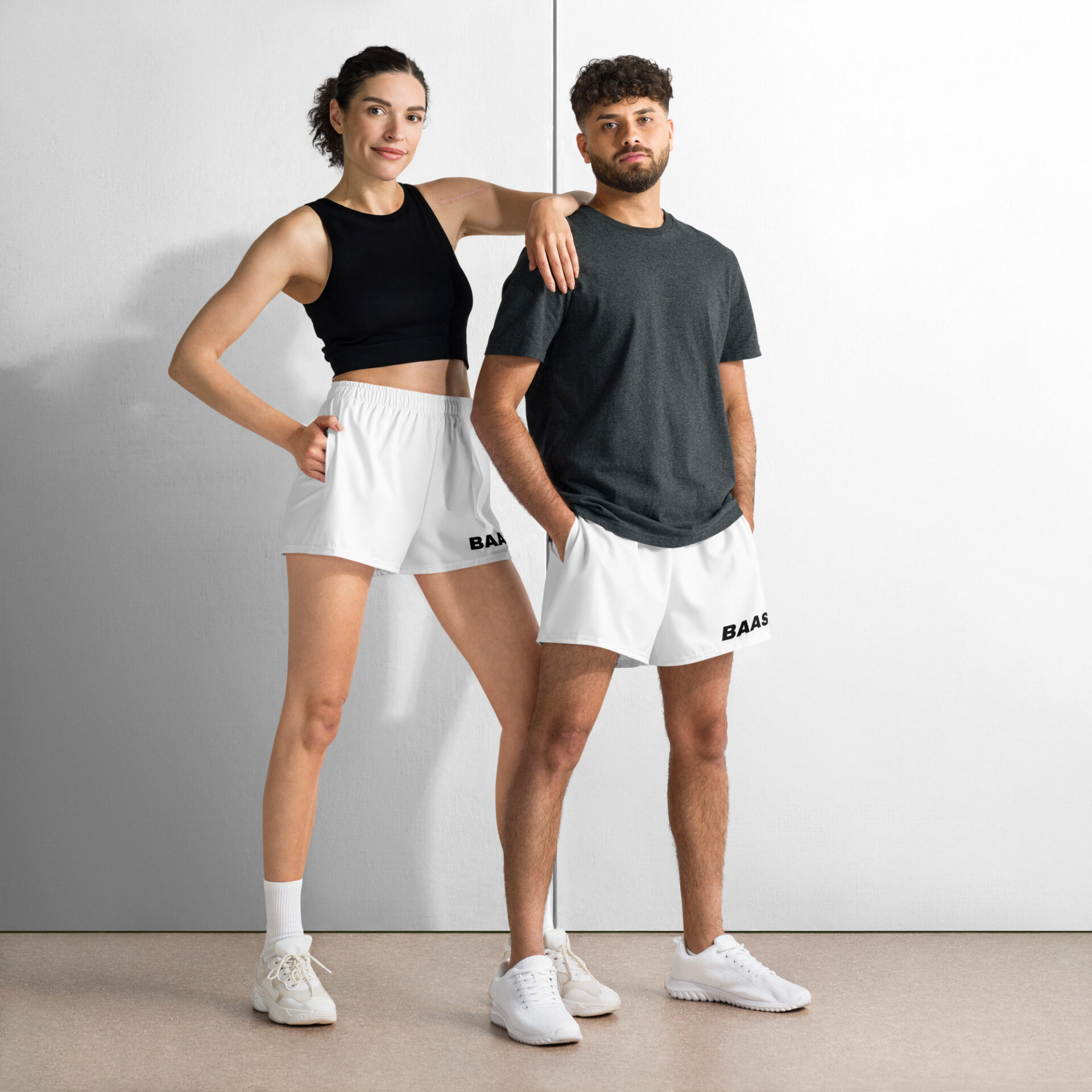 A woman and man stand confidently against a white wall; she in a black sleeveless top and athletic shorts, he in a dark gray T-shirt and BAAS Athletic Shorts. Both complete their look with crisp white sneakers. A woman and man stand confidently against a white wall; she in a black sleeveless top and athletic shorts, he in a dark gray T-shirt and BAAS Athletic Shorts. Both complete their look with crisp white sneakers.