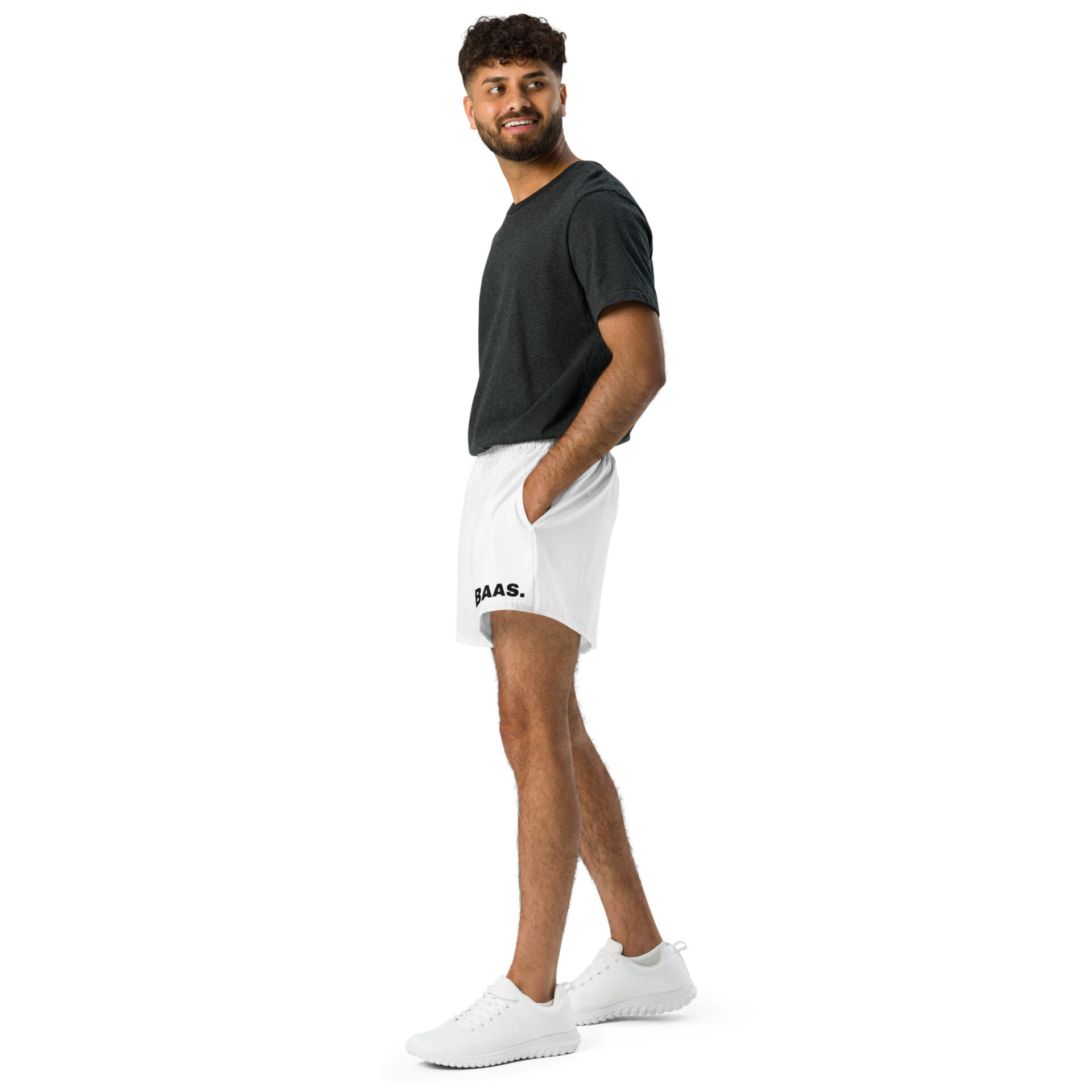 A bearded man smiles in dark gray T-shirt and All-Over Print Unisex Athletic Shorts with BAAS, wearing white sneakers. His hands are in his pockets, and he stands slightly angled against a white background. A bearded man smiles in dark gray T-shirt and All-Over Print Unisex Athletic Shorts with BAAS, wearing white sneakers. His hands are in his pockets, and he stands slightly angled against a white background.