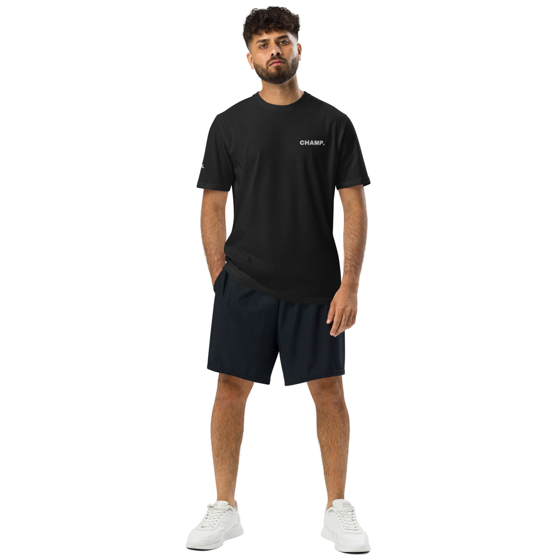 A man with curly hair and a beard stands facing forward, wearing an Under ArmourⓇ athletic t-shirt CHAMP, dark shorts, and white sneakers against a plain white background. His arms are relaxed by his sides. A man with curly hair and a beard stands facing forward, wearing an Under ArmourⓇ athletic t-shirt CHAMP, dark shorts, and white sneakers against a plain white background. His arms are relaxed by his sides.